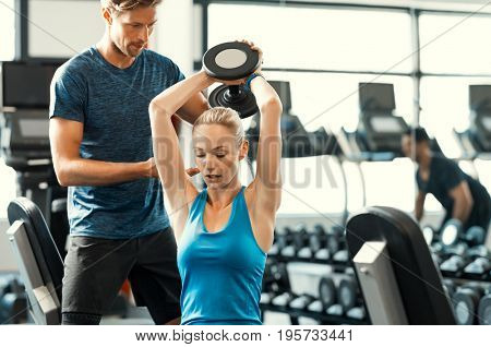 Trainer helping athletic woman at gym. Personal trainer giving weightlifting training to girl in gym. Young woman working out at gym using dumbbells with help of personal trainer.