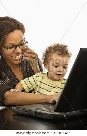 African American mid adult businesswoman at work on laptop and cell phone with toddler son on lap.