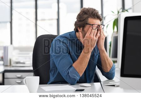 Portrait of an upset businessman at desk in office. Businessman being depressed by working in office. Young stressed business man feeling strain in eyes after working for long hours on computer.