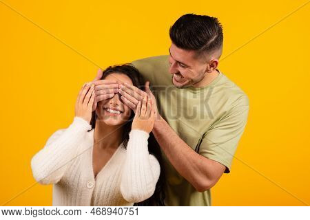 Smiling Millennial Arabic Guy Closes Eyes Of Surprised Lady, Birthday Greeting, Isolated On Orange B