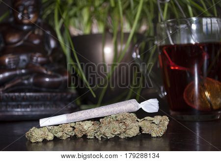 closeup of rolled marijuana weed joint and buds on wooden background with Buddha statuette and cup of rooibos tea