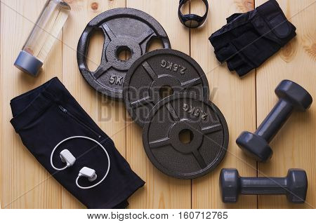 Top view of accessories for fitness in black grey tone. Dumbbells weight plates gloves for gym sport watchmusic player and bottle of water on wooden background. Concept for sport or workout.