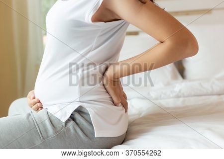 Pregnant Woman Touching Tummy And Suffering From Back Ache Sitting On Bed At Home. Young Attractive 