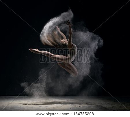 Gymnast in beige body jumping gracefully in dust cloud black background studio shot