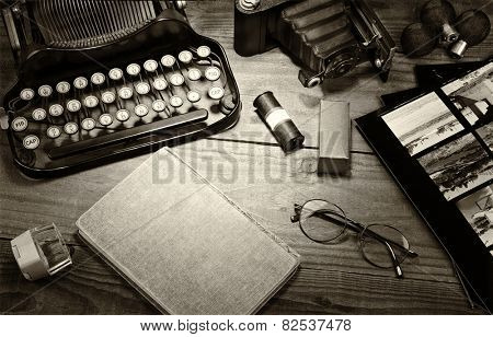 Closeup of a vintage photography still life with typewriter, folding camera, loupe, roll film, flash bulbs, contact prints and book on a wood table. Black and white toned image for a vintage feel. 