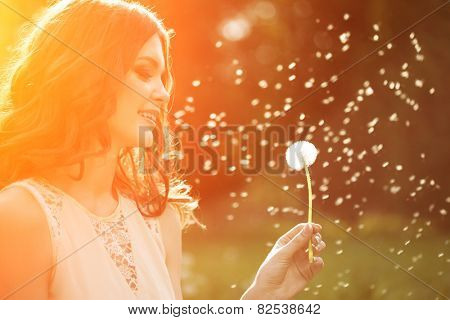 Young spring fashion woman blowing dandelion in spring garden. Springtime. Trendy girl at sunset in spring landscape background. Allergic to pollen of flowers. Spring allergy 