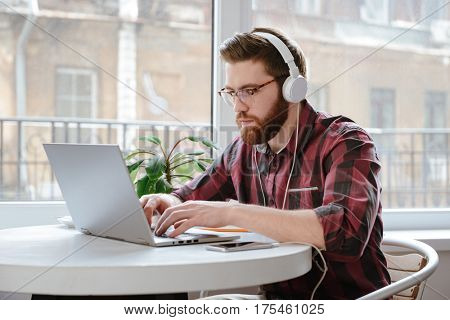 Image of concentrated bearded young man student sitting in cafe while using laptop computer and listening music. Looking at laptop.