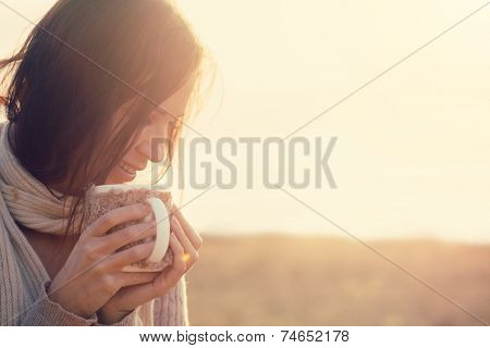 Woman wearing warm knit clothes drinking cup of hot tea or coffee outdoors in sunlight