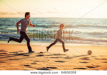 Happy father and son play soccer or football on the beach in sunset light