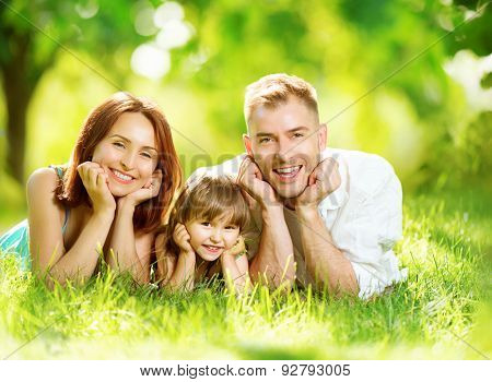 Happy joyful young family father, mother and little daughter having fun outdoors, playing together in summer park. Mom, Dad and kid laughing, lyying on green grass, enjoying nature outside. Sunny day