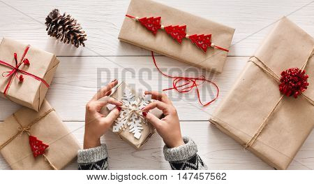 Creative hobby. Woman's hands wrap christmas holiday handmade present in craft paper with twine ribbon. Making bow at xmas gift box, decorated with snowflake on white wooden table, top view.