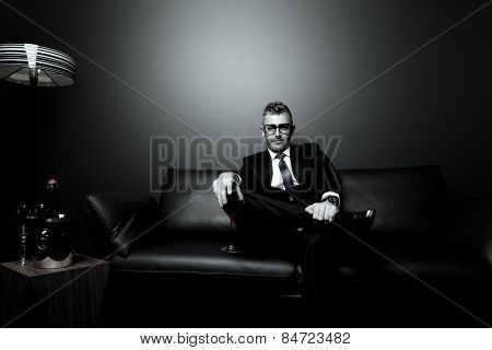 Black-and-white portrait of a handsome mature man in elegant suit drinking red wine. He is sitting on a leather couch in a luxurious interior. 