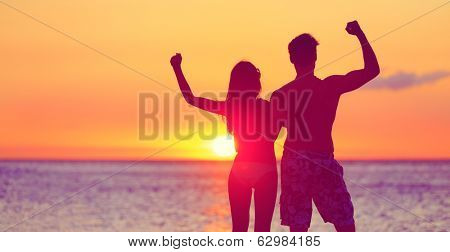 Happy fitness people on beach at sunset flexing showing muscles. Cheering winning couple expression joy and success together embracing. Man and woman on tropical beach.