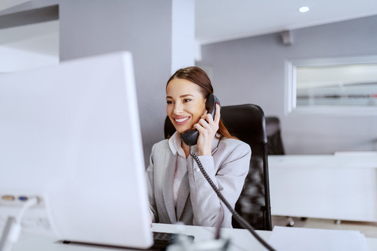 Gorgeous Caucasian businesswoman with long brown hair and in formal wear using computer and talking on the phone. Multitasking concept.