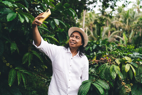 Smiling male farmer using cellphone technology for making selfie photos near coffee bush at own plantation in Indonesia, happy man with mobile device photographing himself at caffeine farmland