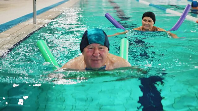 Portrait of smiling happy positive white senior adult man with blue swimming swim cap on his head using foam green pool noodle. Indoor shot. Sports area. High quality 4k footage