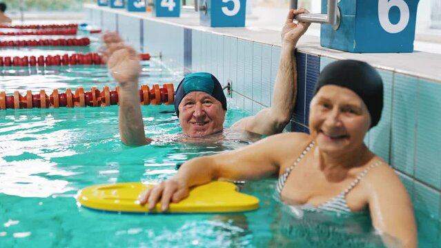 positive energetic caucasian senior couple submerged in turquoise swimming pool water with hands up in the air. Swimming gear and equipment. Looking at camera. High quality 4k footage