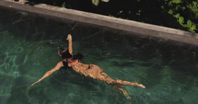 Aerial drone view of a slim young adult sexy woman in black bikini swimming in a luxury tropical resort pool. Slow motion 4k footage