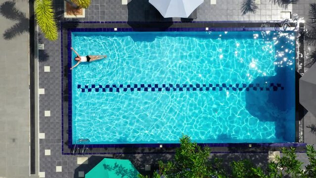 4K footage over swimming pool with relaxing woman in white swimsuit floating on the blue ripples water surface. Holidays, recreation concept.