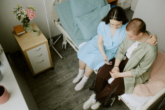 Pregnant woman hugging her friend and showing photos via phone while they sitting on hospital bed