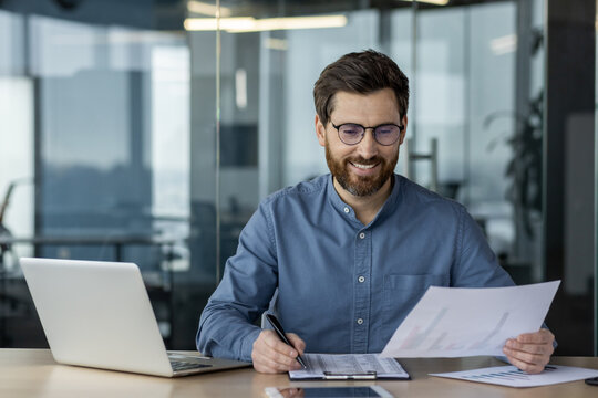 Smiling young man businessman in glasses works in office with laptop and documents, studies data, conducts analysis and examination