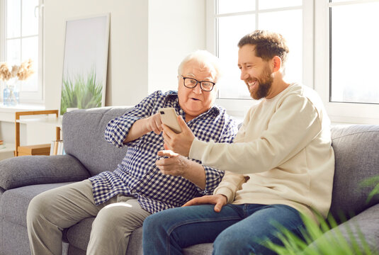 Old father and adult son sitting on sofa looking photos on phone laughter and smiling at whole room. Old father and adult son family moments happiness and love father and son looking photos on phone