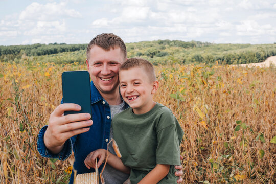 Agronomist, father and son together in agriculture field with soybeans. The man with boy are doing selfie on the phone. Quality control of crops. Front view