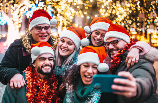 Self portrait of young people wearing santa claus hat celebrating Christmas day outside - Happy multiracial friends group taking selfie under tree having xmas holiday party - Winter holidays concept