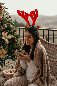 A smiling young woman enjoying a Christmas morning, holding a cup of tea resting on the terrace of a modern barn house in the mountains