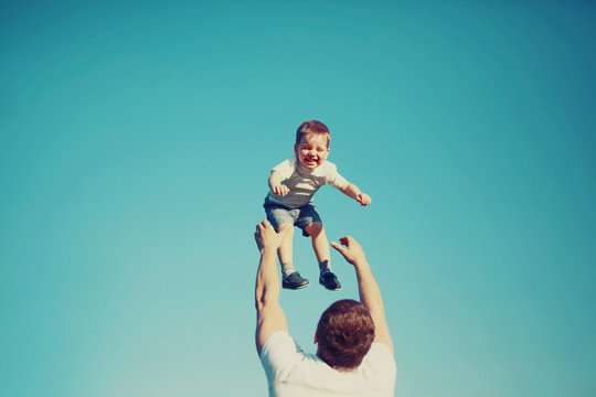 Happy father and son having fun, dad throwing child in the air outdoors, blue sky background, vintage photo 