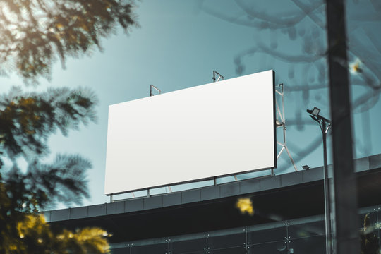 An empty huge poster mockup on the roof of a mall; white template placeholder of an advertising billboard on the rooftop of a modern building framed by trees; blank mock-up of an outdoor info banner