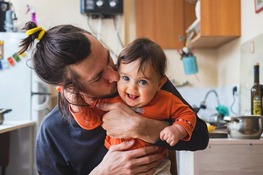 Hipster young father hug and kiss his baby infant boy holding on hands. At home on kitchen background. Lifestyle photo of family care and happiness concept. Middle class. 