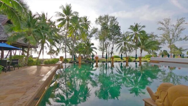 Outdoor swimming pool by ocean beach with reflection on cyan water surface of bright sun light ray shining trough green palm tree leaf on white cloud blue sky background, 4k cinemagraph b-roll footage