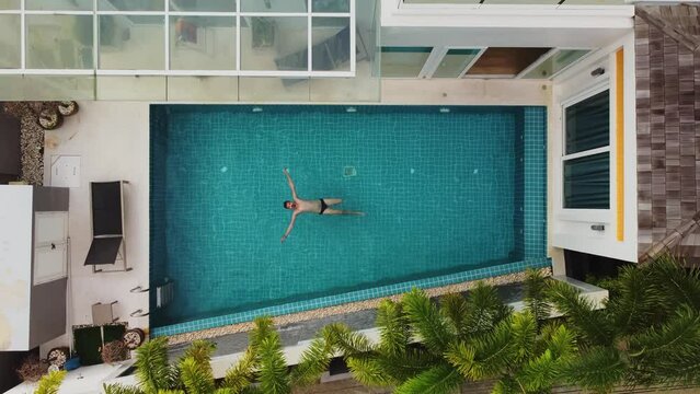 Man is floating on his back in open swimming pool and moving legs in water filmed from above by drone on the backyard of hotel room. Aerial footage of caucasian t tourist relaxing at the poolside