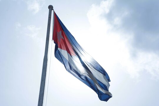 The national flag of Cuba flies up a flagpole on a beautiful summer day with a background of blue sky and white clouds.