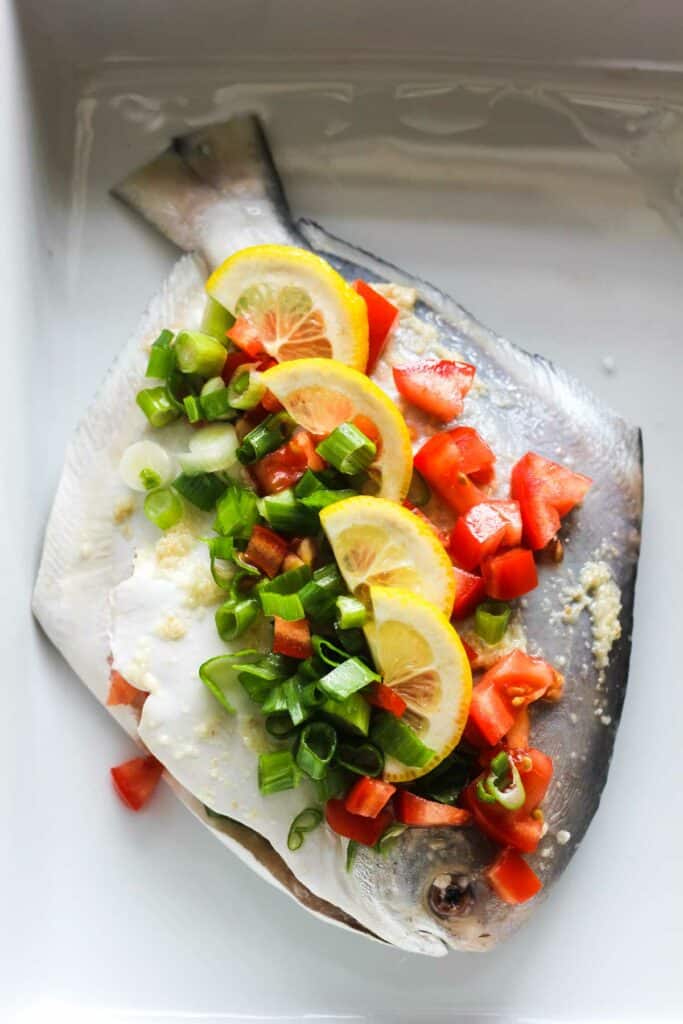 raw pomfret fish covered with tomatoes, green onions and lemon slices