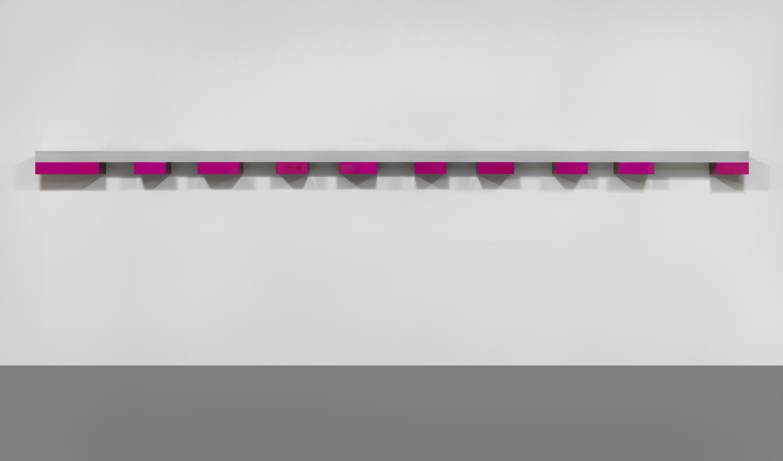 A minimalist art installation featuring a long horizontal shelf with evenly spaced magenta squares that create a repeating pattern against a white wall.