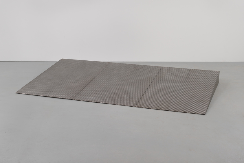 A large, textured metal sheet lies flat on a grey floor against a white wall, with the sheet appearing to be folded or creased in several places to create a three-panel effect.