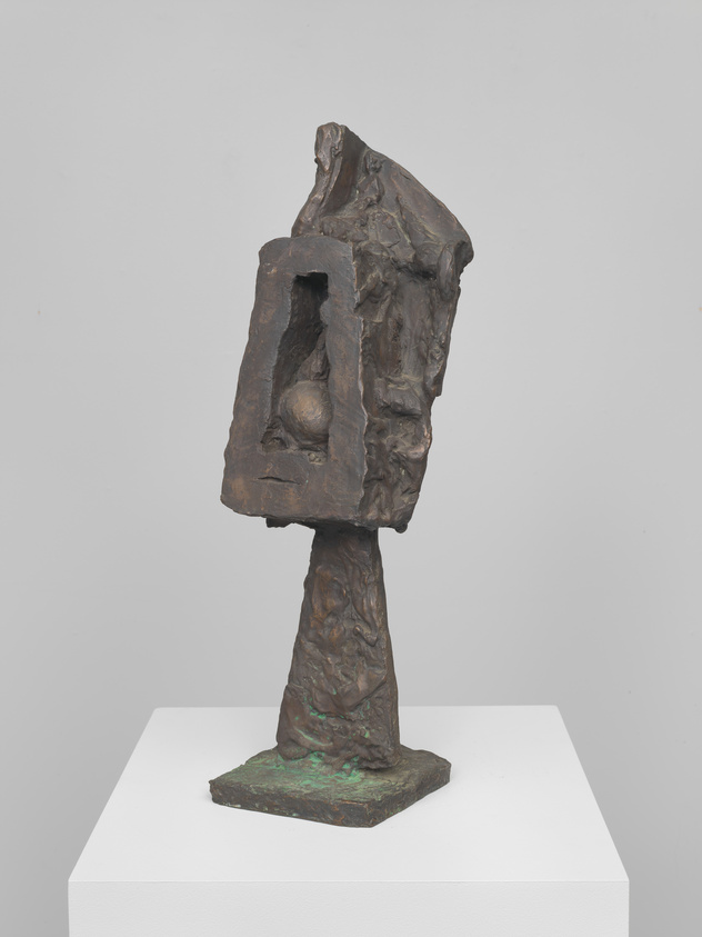 A bronze abstract sculpture with a rough texture, featuring an irregularly shaped head atop a tapered base, displayed on a white pedestal against a light background. The head portion has a prominent hollowed-out section resembling a facial cavity.