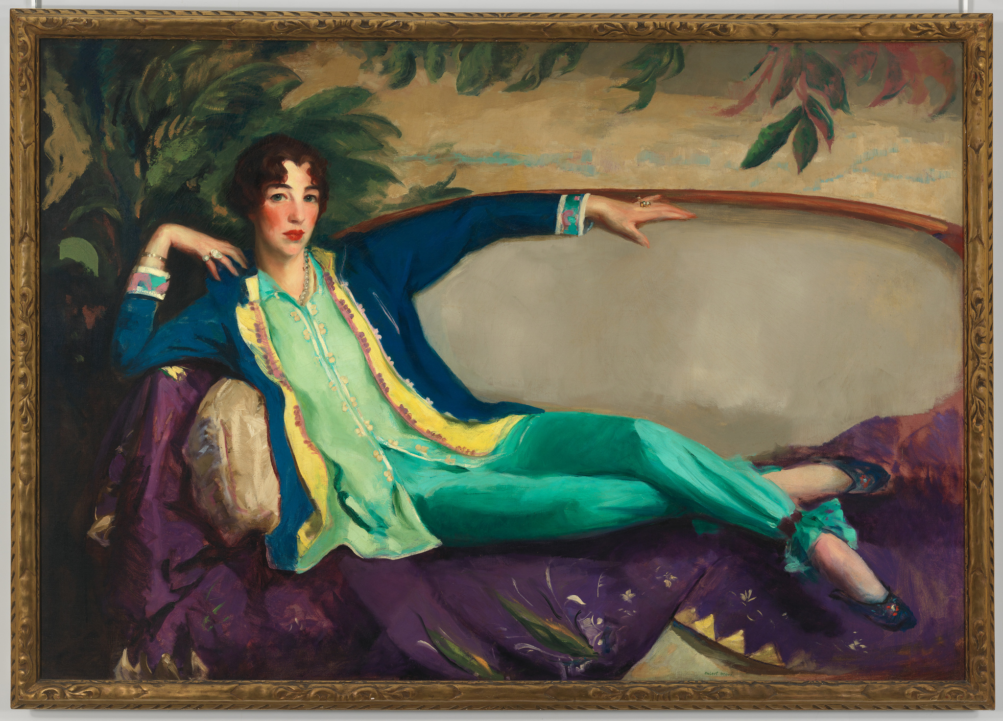 A white woman with short brown hair, rosy cheeks, and dressed in a flowy teal and blue shirt and pants lounges on a sofa while gazing intently.