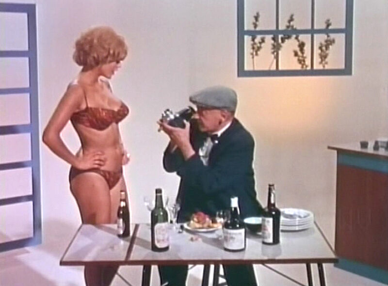 A still from a film. A seated man points a camera at a woman in her underwear.