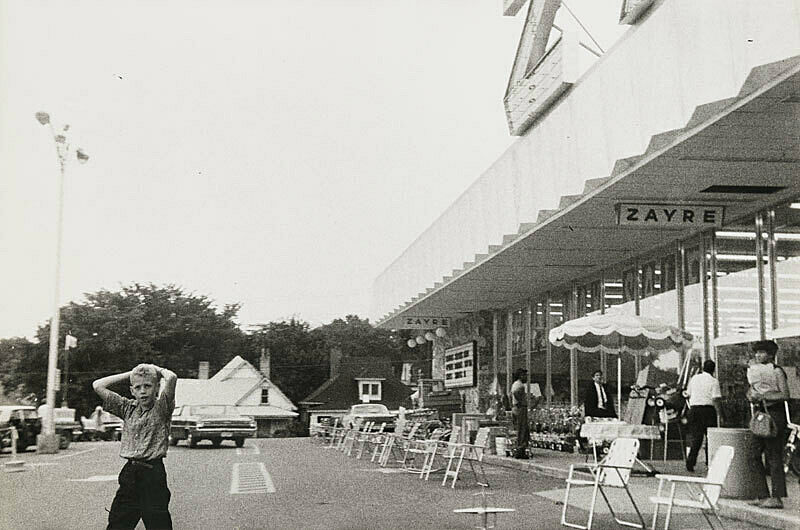 Black and white photograph of parking lot with lawn chairs lining the side walk and a young boy facing camera.