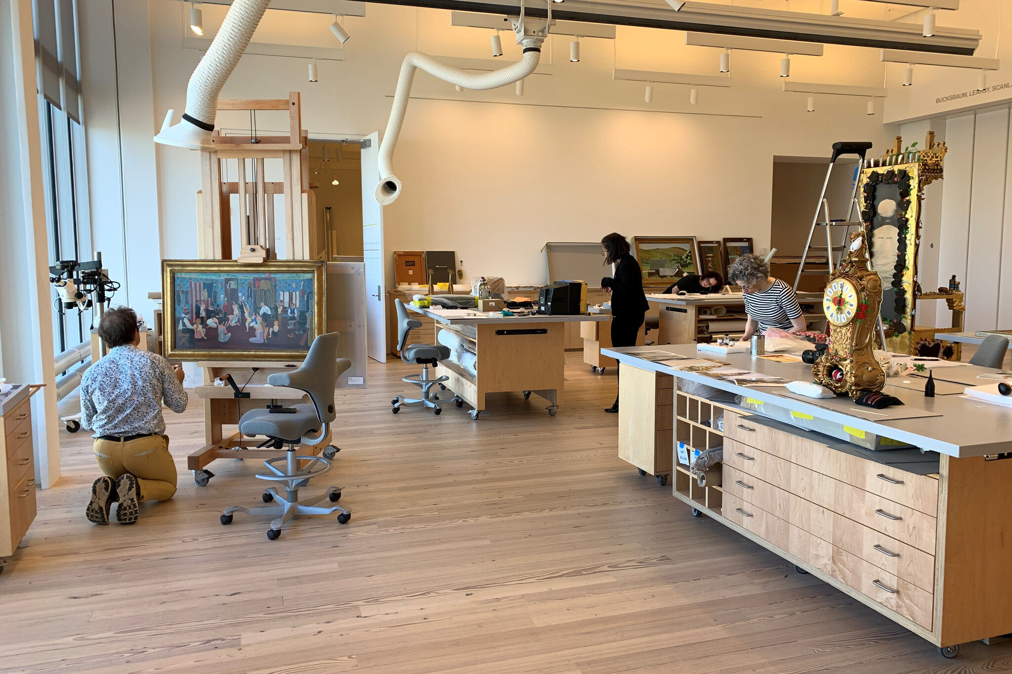 Art conservators work on various pieces in a well-lit studio, including a large painting and an ornate clock.