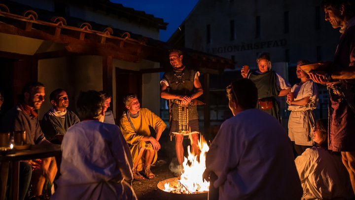 Römisch gekleidete Männer und Frauen nachts an einem Lagerfeuer in historischer Kulisse