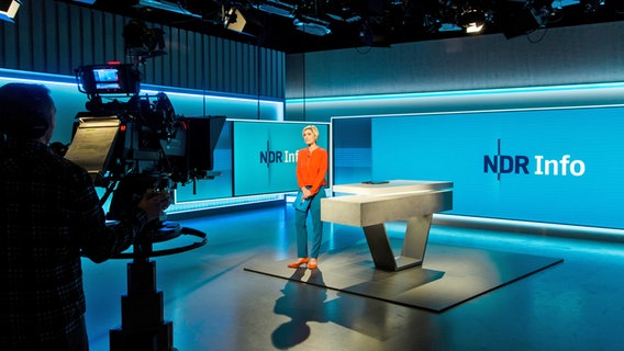 Blick ins NDR Info Fernseh-Studio. Zu sehen: ein Kameramann an der Kamera von hinten filmt Moderation Susanne Stichler, die am Moderationstisch steht. © NDR/Hendrik Lüders Foto: Hendrik Lüders