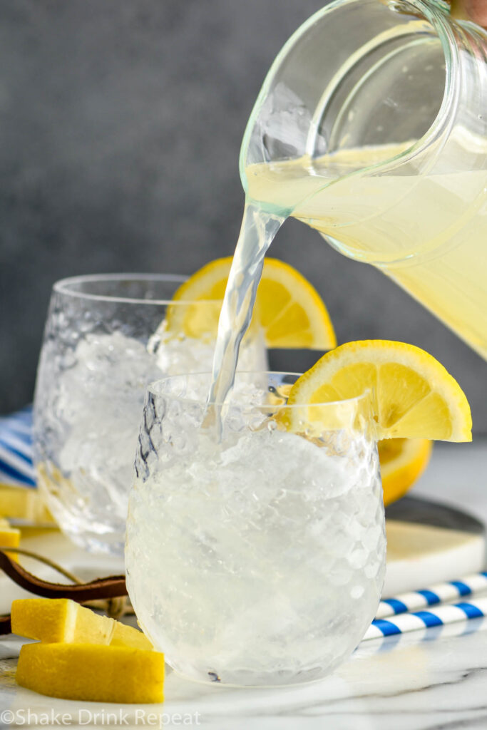 pouring pitcher of lemonade into a glass filled with ice and vodka to make a vodka lemonade with slices of lemon for garnish