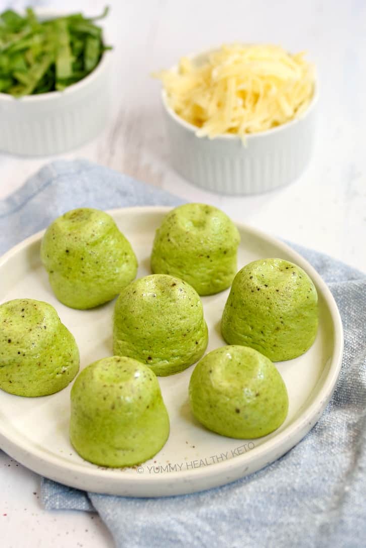 A white plate holding 7 Instant Pot Keto Egg Bites, sitting on a pale blue napkin with a small bowl of shredded cheese and a small bowl of chopped spinach in the background.